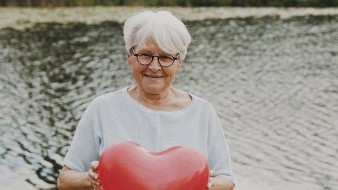 Senior woman holding heart shaped balloon near the river. Love and care concept. High quality 4k footage