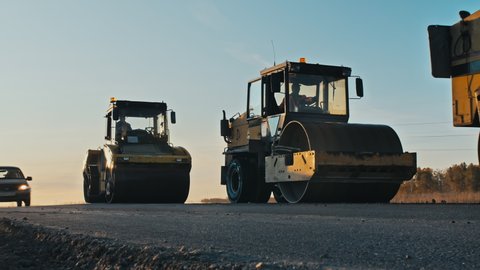 Road surface repair. Construction of a new road. Three rollers level and compact the asphalt at sunset, cars pass by. A layer of freshly laid asphalt in the rays of the setting sun.