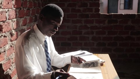 A slow motion side view shot of a young African male in a school uniform as he studies his books in an old run down brick school in Africa.