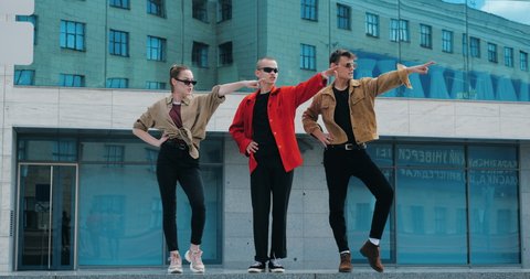 Three performer street dancers rehearse outdoor dance near blue building. Group teenagers wears stylishly fashionable shirts black sunglasses, stands in line, make synchronized movements with hands