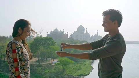 A smiling man surprises girlfriend with a wedding on romantic holidays. A woman gets excited after a boyfriend proposes her for marriage against beautiful view of the Taj mahal in the background