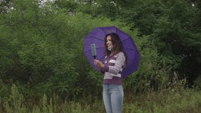 A young Caucasian woman in a coronavirus mask is vlogging in the forest. A woman under an umbrella in the forest holding an umbrella communicates on a video call using a smartphone.