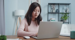 Charming Asian woman sitting at table and working in home office room. Concentrated female worker typing, shutting laptop computer and looking at camera. Self-isolation, occupation, job.
