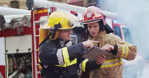 Medium plan of two firefighters discussing rescue plan looking at tablet while standing in helmets near a fire van. Smoke from the fire covers rescuers and a fire truck.