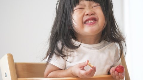 Asian preschool toddler girl eats cherry tomato with hand and becomes smile. Safety and healthy food ingredients. A daughter sitting the child chair
