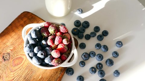 White Ceramic Bowl with Fresh Blueberry and Freshly Frozen Strawberry Standing on Wooden Rustic Cutting Board on Table with Glass Jar of Milk. Healthy Vegetarian Food. Top view. Berries Breakfast.