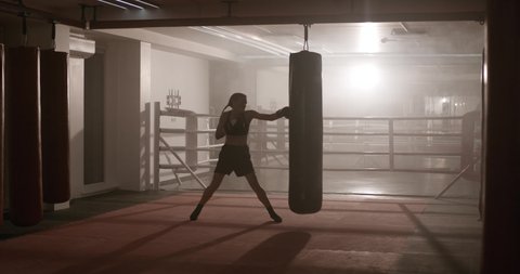 Sports motivation, kickboxing, woman fighter trains his punches, beats a punching bag, training day in the boxing gym, back lighting, 4k Slow Motion.