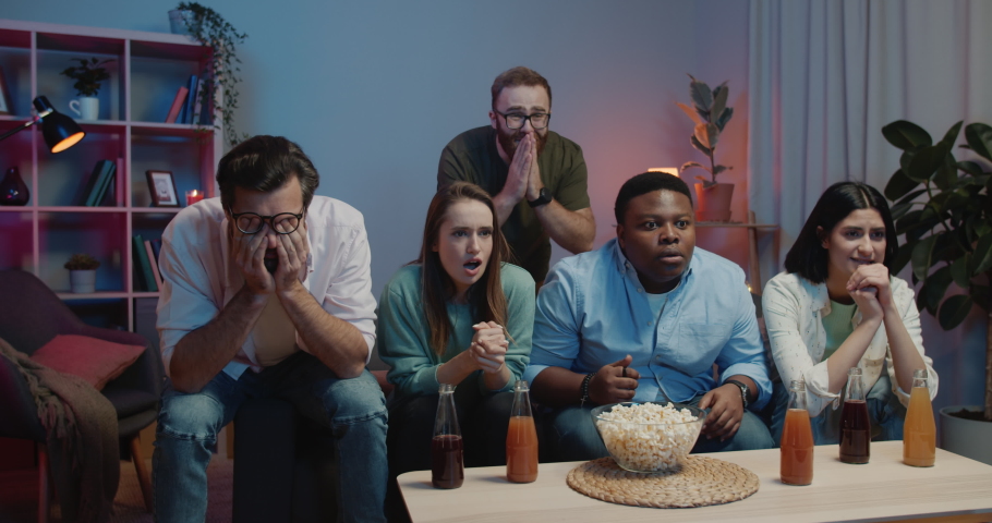Friends watching sports on TV and making enthusiastic face expression. Thrilled group of youth sitting on sofa and supporting sport team with drinks and snacks. | Shutterstock HD Video #1058760694