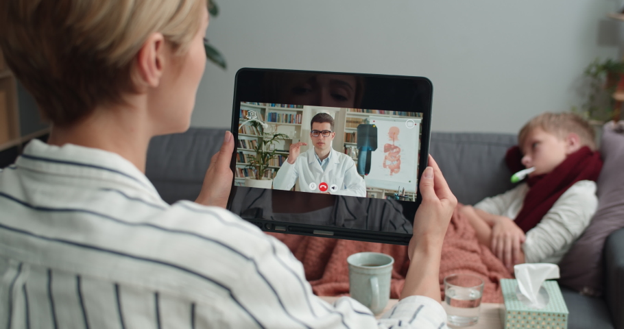 Backside view of mother holding tablet and having online medical consultation with male doctor while her ill son lying on sofa at home. Concept of illness and telehealth. | Shutterstock HD Video #1058760715