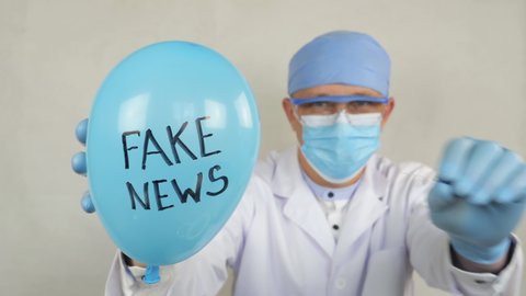 Fake News. Doctor holds a balloon with the inscription Fake News. Doctor in a medical gown and a protective mask punch balloon with a medical needle.