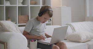 preschooler boy is learning to draw at home, listening music and training video through headphones by laptop