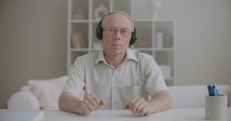 aged man with headphones is looking at camera, greeting online audience and starting learning webinar Royalty-Free Stock Footage #1058763820