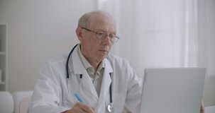 elderly professor of medicine is viewing webinar by laptop in office of clinic and making notes, e-learning for health professionals
