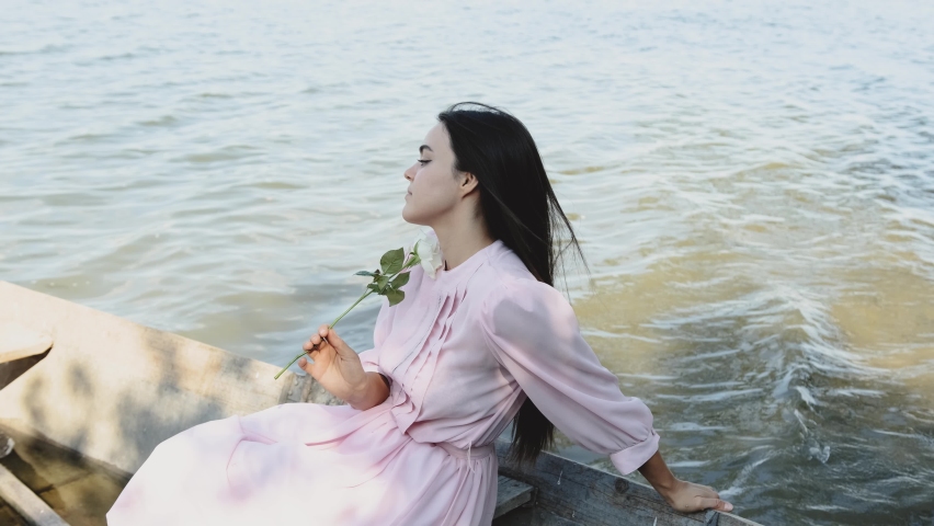 A girl dressed in a pink dress sits in a boat and admires the view, the girl holds a flower in her hand.