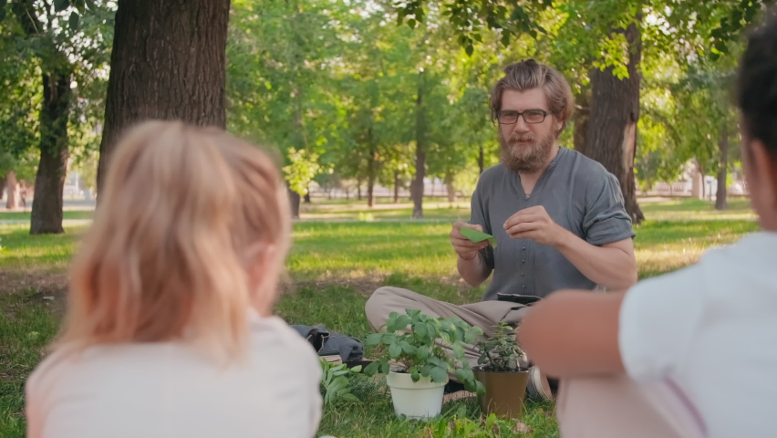 Tracking shot of bearded male biology teacher in glasses sitting cross-legged on blanket in park and teaching curious kids about nature