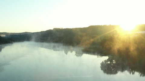 Early morning mist floating over calm water, lake at hilly landscape. Aerial view of slowly floating vapor, steam over lake at dawn time, sunrise with sun rays.