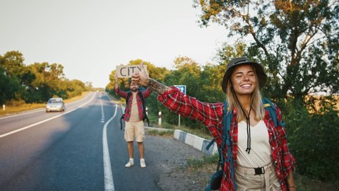 Young millennial woman hitchhiking on road with boyfriend who holding sing City
