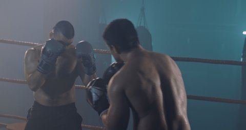 Caucasian young male shirtless boxers punching each other in dark ring at training. Strong sweaty men exercising and boxing in darkness. Shirtless sportsmen in gloves fighting.