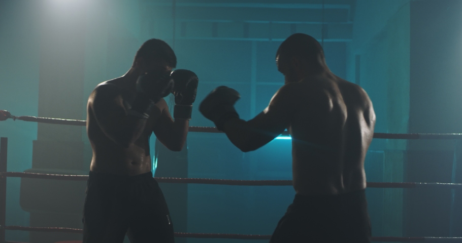 Caucasian two professional young shirtless male boxers fighting at dark boxing ring. Men kickboxers kickboxing in darkness at training. Fight sport concept.