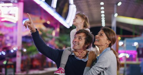 Authentic shot of a happy smiling family is having fun together in amusement park with luna park lights at night.