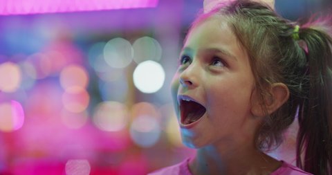 Authentic close up shot of a happy excited smiling little girl is having fun in amusement park with luna park lights at night.
