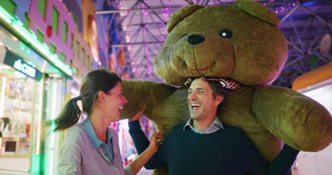 Authentic shot of a happy carefree smiling couple in love is having fun with giant peluche eddy bear won with play fair shooting games in amusement park with luna park lights at night.