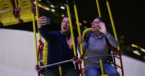 A happy carefree smiling couple in love is having fun to make selfie or video call to friends or family while riding chain swing carousel together in amusement park at night.