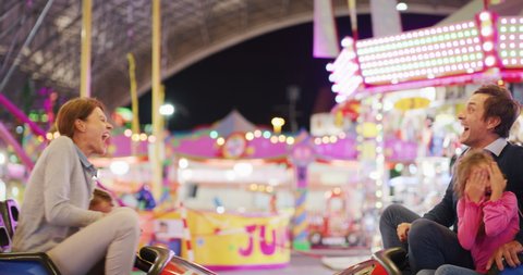 Authentic shot of a happy smiling family is having fun to drive bumper cars at fun fair in amusement park with luna park lights at night.