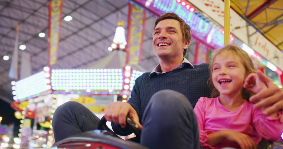 Authentic shot of a happy smiling father and little daughter are having fun to drive a bumper car together at fun fair in amusement park with luna park lights at night. Royalty-Free Stock Footage #1058792251