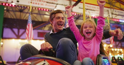 Authentic shot of a happy smiling father and little daughter are having fun to drive a bumper car together at fun fair in amusement park with luna park lights at night.