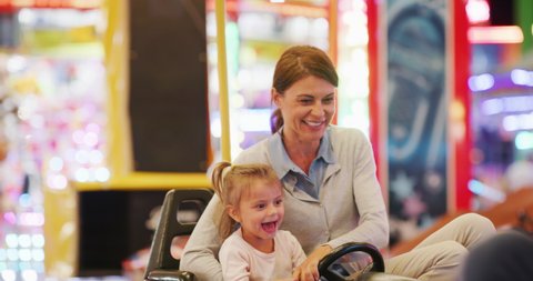 Authentic shot of a happy smiling mother and little daughter are having fun to drive a bumper car together at fun fair in amusement park with luna park lights at night.