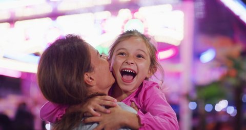 Authentic close up shot of a happy smiling daughter is running to give an affective hug and kiss to her mother while having fun together in amusement park with luna park lights at night.