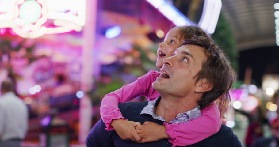 Authentic shot of a happy smiling father carrying his little daughter on a shoulders having fun together in amusement park with luna park lights at night. | Shutterstock HD Video #1058793082