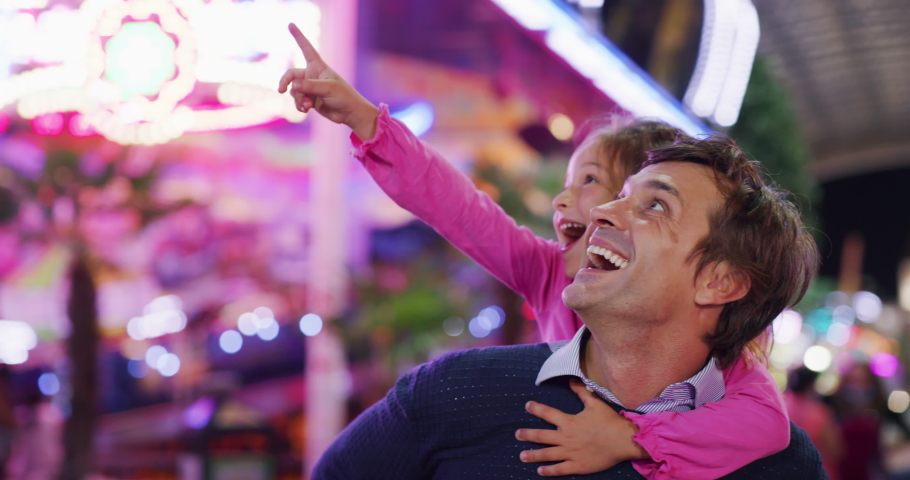 Authentic shot of a happy smiling father carrying his little daughter on a shoulders having fun together in amusement park with luna park lights at night. | Shutterstock HD Video #1058793082