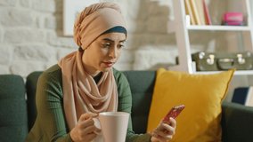 Beautiful Muslim woman reading a message on her smart phone while sitting at her home. Young woman is drinking coffee while reading a message.Close up and slow motion video.
