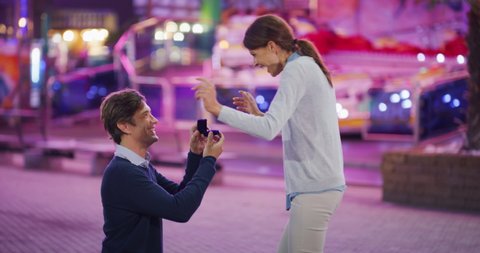 Authentic shot of young handsome man is making a surprise proposal of marriage to his beloved woman in amusement park with luna park lights at night. 