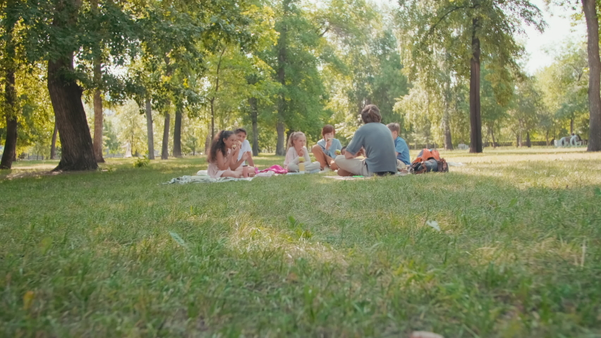 Dolly-in ground level shot of male teacher with magnifying glass sitting on blanket on grass in park and talking to group of schoolchildren eating lunch and listening to him attentively