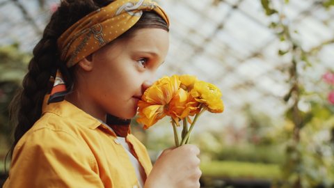 Selective focus shot of beautiful little girl smelling yellow flowers, looking at camera and smiling while standing in greenhouse farm