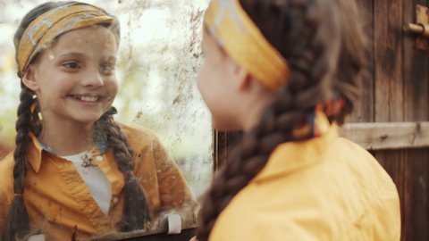 Cute little girl in yellow headband and shirt walking along wooden wall in greenhouse farm, looking at her reflection in rusty mirror and smiling