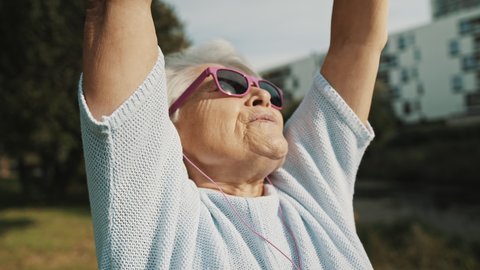 Cool grandmother with pink sunglasses and headphones stretching in the park. Recreation in old age. High quality 4k footage Stockvideo