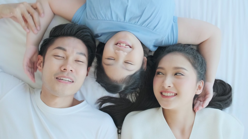 Happy Asian family mom dad and daughter child laying laugh smile on white soft bed in bedroom relax casual leisure weekend activity, top view handheld shot | Shutterstock HD Video #1058803159