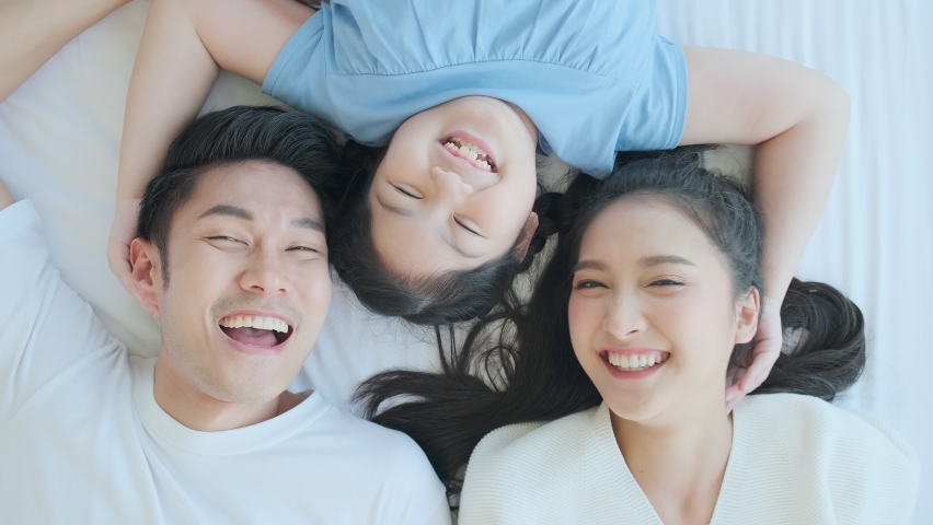 Happy Asian family mom dad and daughter child laying laugh smile on white soft bed in bedroom relax casual leisure weekend activity, top view handheld shot | Shutterstock HD Video #1058803159