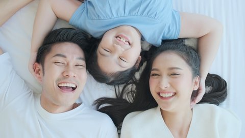 Happy Asian family mom dad and daughter child laying laugh smile on white soft bed in bedroom relax casual leisure weekend activity, top view handheld shot