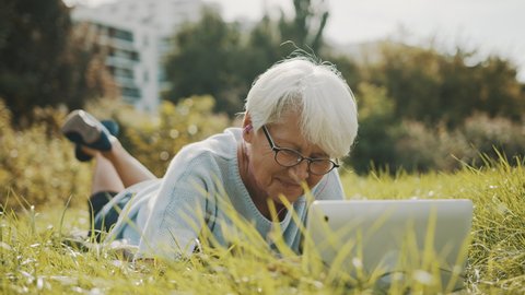 senior retired woman using laptop in the nature while lying on the grass. High quality 4k footage