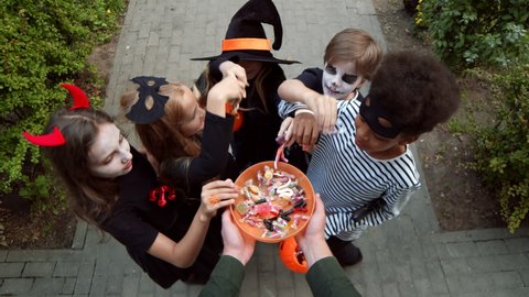 First person view of five diverse kids wearing cool Halloween costumes are talking different lollipops and candies out of bowl unrecognizable person holding in hands