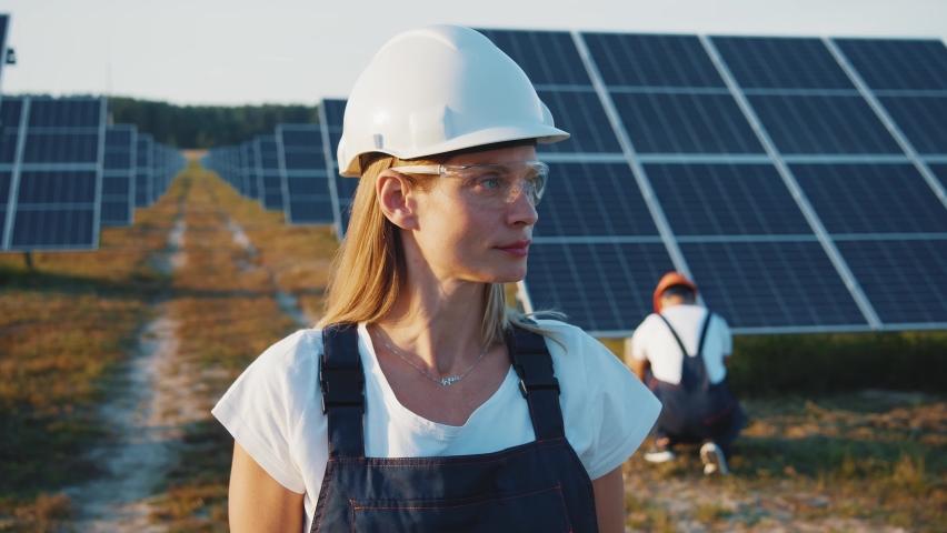 Portrait of nice-looking cheerful female blonde engineer in uniform standing by solar batteries. Woman worker cooperating with colleague on ecological solar farm. Royalty-Free Stock Footage #1058809258