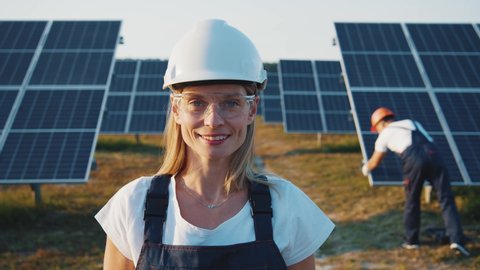 Portrait of nice-looking cheerful female blonde engineer in uniform standing by solar batteries. Woman worker cooperating with colleague on ecological solar farm.