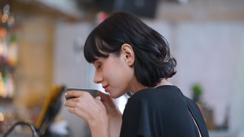 Close up and slow motion shot of short dark hair European woman with white skin sits and enjoys smelling a dark grey cup of coffee and feeling appreciate at coffee bar in cafe restaurant.