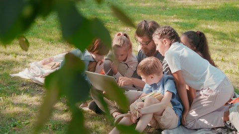 Slowmo tracking shot of curious schoolchildren and their male bearded teacher looking at laptop while learning in park on sunny summer day