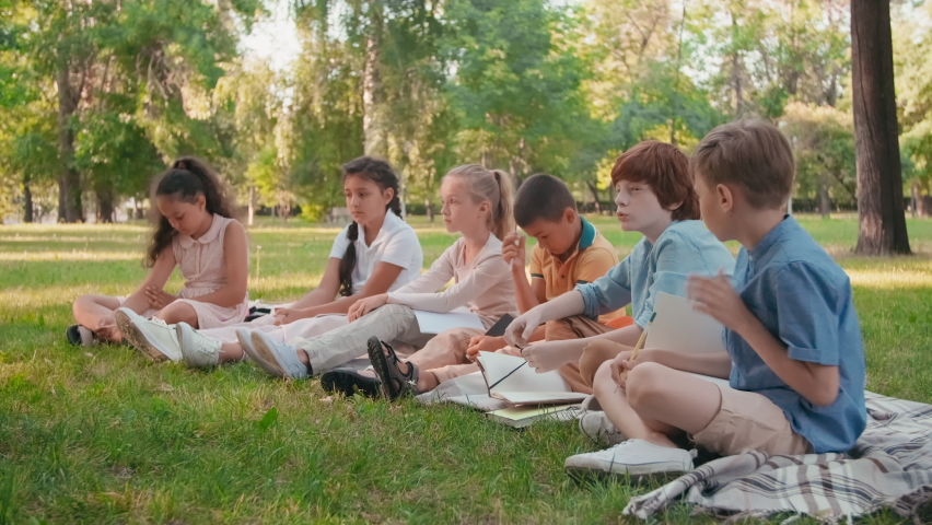 Medium shot of schoolchildren sitting on blankets in park on sunny summer day and learning